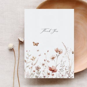 Puede incluir: Una tarjeta blanca con el texto "Gracias" en caligraf&iacute;a negra. La tarjeta presenta una ilustraci&oacute;n de acuarela de un campo de flores silvestres y mariposas en tonos marrones, rosas y verdes.