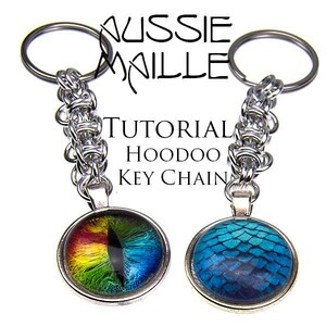 Peut inclure: Deux porte-clés argentés avec une chaîne argentée et un charme rond. Les breloques sont un motif d'écailles de dragon bleu et vert et un motif d'œil arc-en-ciel. Le texte "TUTORIAL HOODOO KEY CHAIN" est visible sur l'image.