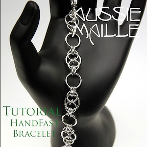 Op de afbeelding: Een zilveren kettingarmband gemaakt met een handfast-patroon. De armband bevindt zich op een zwarte, handvormige displaystandaard. De tekst "TUTORIAL HANDFAST BRACELET" bevindt zich onder de armband. De tekst "Aussie Maille" bevindt zich boven de armband.