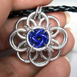 May include: A silver chainmail flower pendant with a blue center. The pendant is attached to a black leather cord.