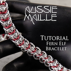 May include: A silver and red chainmail bracelet, known as a "Fern Elf" bracelet, is shown on a black surface. The bracelet is made of small, interlocking metal rings. The text "Aussie Maille Tutorial Fern Elf Bracelet" is visible in the image.