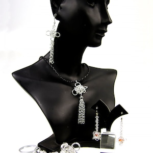 May include: A silver chainmaille necklace, earrings, and keychain with a tassel pendant. The necklace and earrings feature a silver knot design.