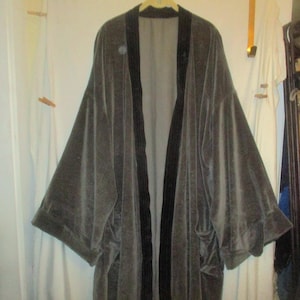 May include: A gray velvet robe with black trim hangs on a wooden hanger. The robe has wide sleeves and a long, open front. The fabric appears soft and drapes loosely. The background includes a white door and a clothing rack.