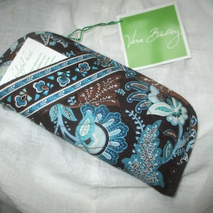 以下が含まれることがあります： 茶色、ティール、白の色合いのフローラルデザインの柄入りメガネケース。 ケースは閉じられており、「Vera Bradley」と書かれたタグが付いています。 ケース内には小さなカードが見えます。