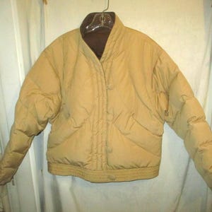 May include: A tan puffer jacket with a brown collar and lining, hanging on a hanger. The jacket has a quilted design, button closures, and elastic cuffs. The jacket is a neutral color.