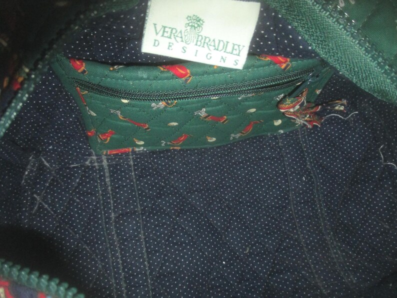 May include: The interior of a quilted bag featuring a dark blue fabric with white polka dots. A green quilted zippered pocket with a golf-themed print is attached, and a white rectangular tag reads Vera Bradley Designs in green text.
