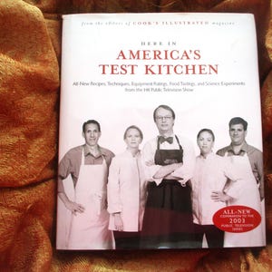 May include: A white ceramic mug with a black handle and the text "But First, Coffee" in a bold, black font. The mug is set against a blurred background, suggesting a cozy setting.