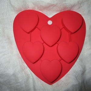 Könnte beinhalten: Eine rote Silikonform in Herzform mit sechs herzförmigen Vertiefungen. Die Form hat oben ein kleines Loch zum Aufhängen. Die Form ist für die Herstellung von herzförmigen Leckereien oder Dekorationen konzipiert. Die Form befindet sich auf einer weißen, strukturierten Oberfläche.