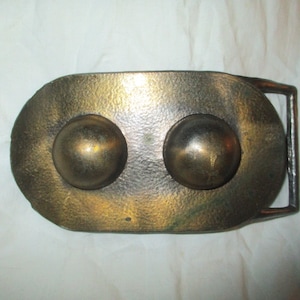 May include: A vintage metal belt buckle with a textured, oval-shaped face. Two large, spherical accents are centered on the face. The buckle has a bronze and gold finish, with a rectangular bar for the belt strap.