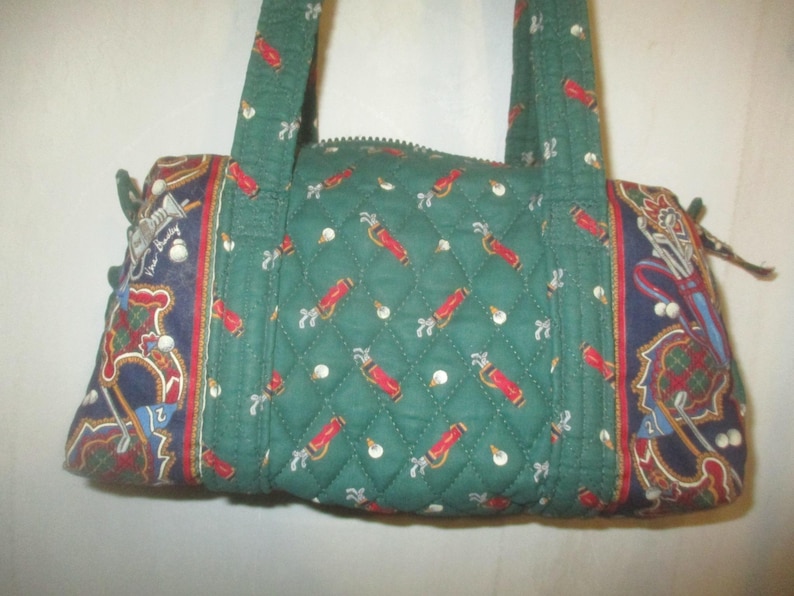 May include: A quilted green handbag featuring a repeating pattern of red golf bags and white golf balls. The side panels are dark blue with a detailed print of golf clubs, bags, and plaid accents. It includes two fabric handles and a zipper.