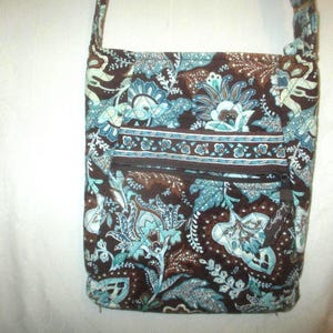 May include: A rectangular crossbody bag featuring a dark brown background with a repeating light blue and white floral paisley pattern. The bag includes a horizontal zippered front pocket with a matching patterned fabric trim and a shoulder strap.