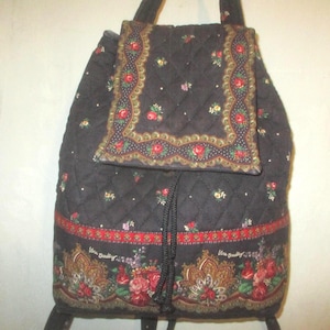 May include: A black quilted backpack featuring a floral pattern with red roses and green leaves. The bag has a drawstring closure, a top flap with a decorative border, and a bottom panel with a detailed floral print and the Vera Bradley logo.