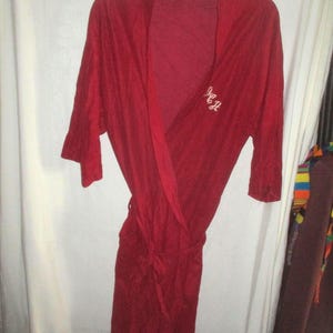 May include: A deep red robe with a tie waist and long sleeves hangs on a hanger. The robe has a wrap-around design and features embroidered initials on the front. The fabric appears soft and lightweight, suitable for lounging.