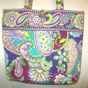 May include: Quilted tote bag with a vibrant paisley floral pattern in shades of purple, teal, green, and pink. Features a wooden toggle closure and matching handles. The Vera Bradley logo is visible.