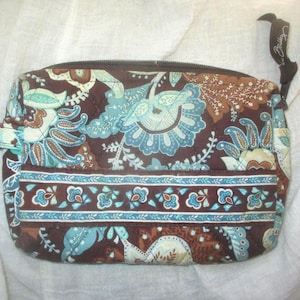 May include: A rectangular quilted cosmetic bag featuring a dark brown background with a light blue, cream, and tan floral paisley pattern. The bag includes a horizontal band across the front, a top zipper closure, and a small fabric side tab.