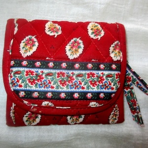 May include: A quilted red wallet featuring a repeating pattern of white and green leaves with small yellow accents. A horizontal band across the center displays a floral motif in red, green, and blue. The wallet includes a fabric zipper pull.