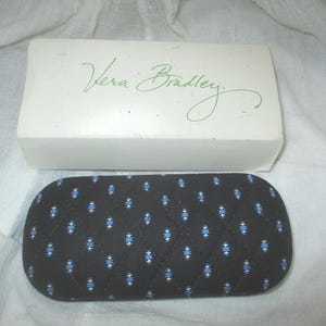 May include: A white Vera Bradley box with the brand name in green script sits above a black, quilted eyeglasses case. The case has a pattern of blue and white geometric shapes.