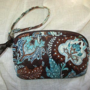 May include: A quilted fabric wristlet pouch featuring a brown background with a repeating paisley pattern in shades of teal, light blue, and white. The pouch has a top zipper closure with a brown ribbon tag and a detachable matching wrist strap.