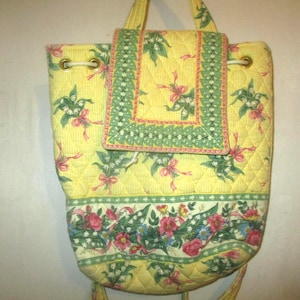 May include: A quilted yellow backpack featuring a floral pattern with green leaves, pink ribbons, and white flowers. The bag has a drawstring closure, a top flap with a green border, and a horizontal floral band across the lower section.