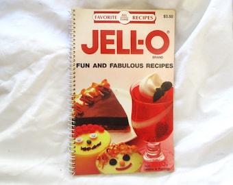 Op de afbeelding: Een spiraalgebonden kookboek getiteld "JELL-O FUN AND FABULOUS RECIPES". De omslag toont het Jell-O-merklogo en afbeeldingen van desserts, waaronder een stuk chocoladetaart en rode gelei.