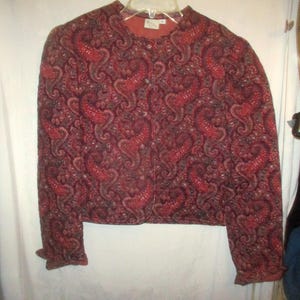 May include: A long-sleeved jacket featuring a dark red and black paisley pattern throughout. The garment has a round neckline, a front button closure, and a quilted texture. The jacket is displayed on a clear plastic hanger against a white background.