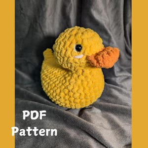 Könnte beinhalten: Ein gehäkeltes gelbes Gummibad-Entchen mit orangefarbenem Schnabel und schwarzen Augen. Die Ente sitzt auf einer grauen, flauschigen Oberfläche. PDF-Muster.