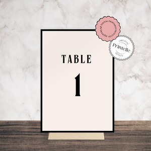 May include: A table number card with a black border. The card has a cream-colored background with the word "TABLE" and the number "1" in a bold, black font. The card is displayed on a wooden stand. Pink and white circular stickers are in the upper right corner.