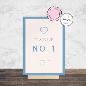 May include: A table number card with a light blue border and text. The card reads "TABLE NO. 1" in a blue font, with the names "LEIGHTON & AUSTIN" below. A crest with the letters "L & A" is at the top.