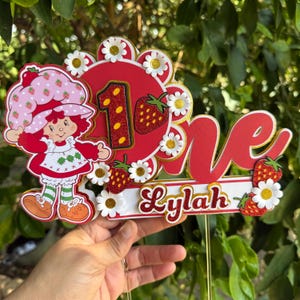 May include: A red and white cake topper featuring a cartoon strawberry shortcake character, the number one, and the name "Lylah". The topper is decorated with white daisies and red strawberries.