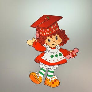 May include: A cartoon character with curly reddish-brown hair wearing a red graduation cap decorated with small strawberries, a red dress with a white apron featuring green leaf patterns, white ruffled bloomers, green striped socks, and orange shoes.