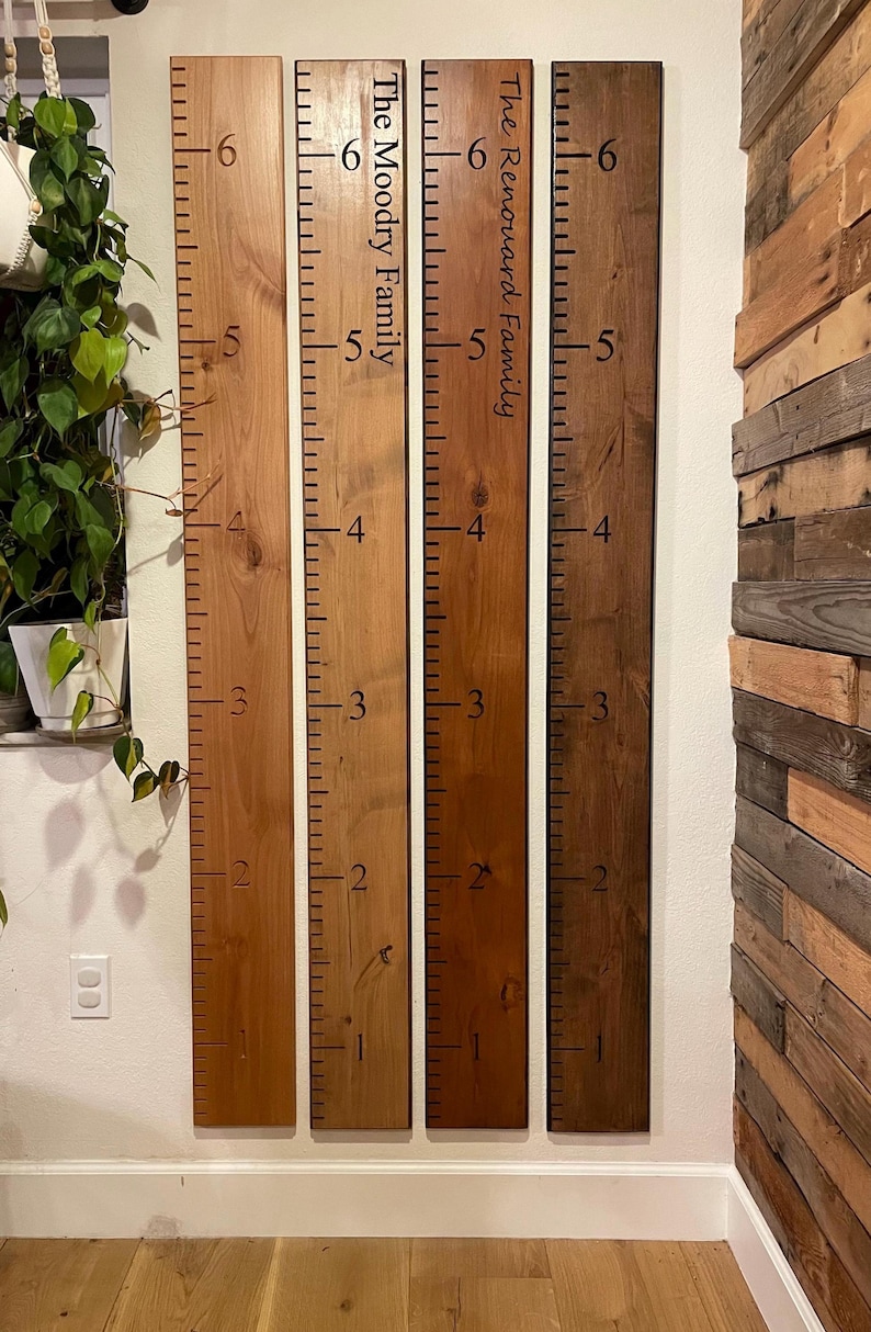 May include: Four vertical wooden growth charts in varying shades of brown, each featuring black measurement markings from one to six feet. Two boards include personalized text reading The Moody Family and The Renouard Family in black ink.