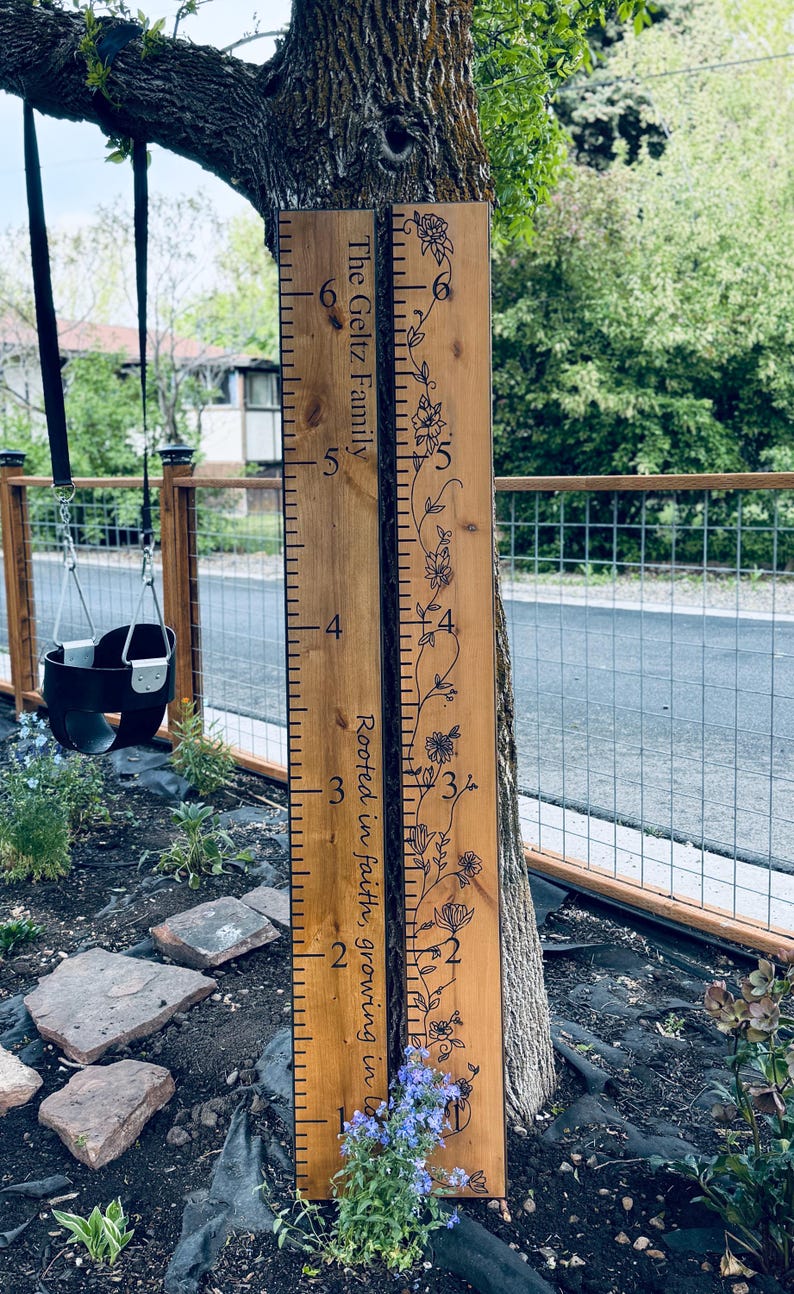 Op de afbeelding: Twee verticale houten groeimeters met zwarte markeringen. Het linkerbord bevat de tekst The Geltz Family en Rooted in faith, growing in love. Het rechterbord toont een zwarte bloemrankillustratie. Beide borden tonen de cijfers 1 tot en met 6.