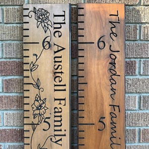 May include: Two vertical wooden growth charts with dark brown stained finishes. The left board features a floral vine engraving and the text The Austell Family. The right board displays the text The Jordan Family. Both include black measurement markings.