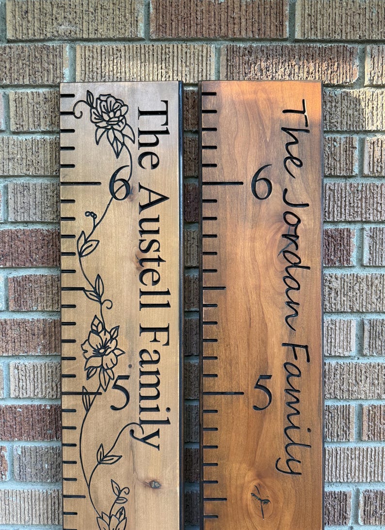 May include: Two vertical wooden growth charts with dark brown stained finishes. The left board features a floral vine engraving and the text The Austell Family. The right board displays the text The Jordan Family. Both include black measurement markings.