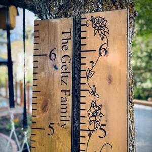 Op de afbeelding: Twee verticale houten groeimeters met zwarte markeringen en cijfers. Het linkerbord bevat de tekst The Geltz Family. Het rechterbord toont een zwarte lijntekening van een klimmende bloemenrank met bladeren en bloemen.