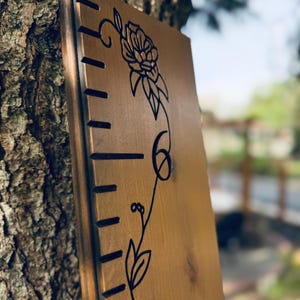 Op de afbeelding: Een verticaal houten groeimeterbord met een gladde, lichtbruine afwerking. Het bord is voorzien van zwarte, lasergegraveerde markeringen langs de linkerrand en een decoratieve zwarte lijntekening van een bloeiende bloemenrank in het midden.