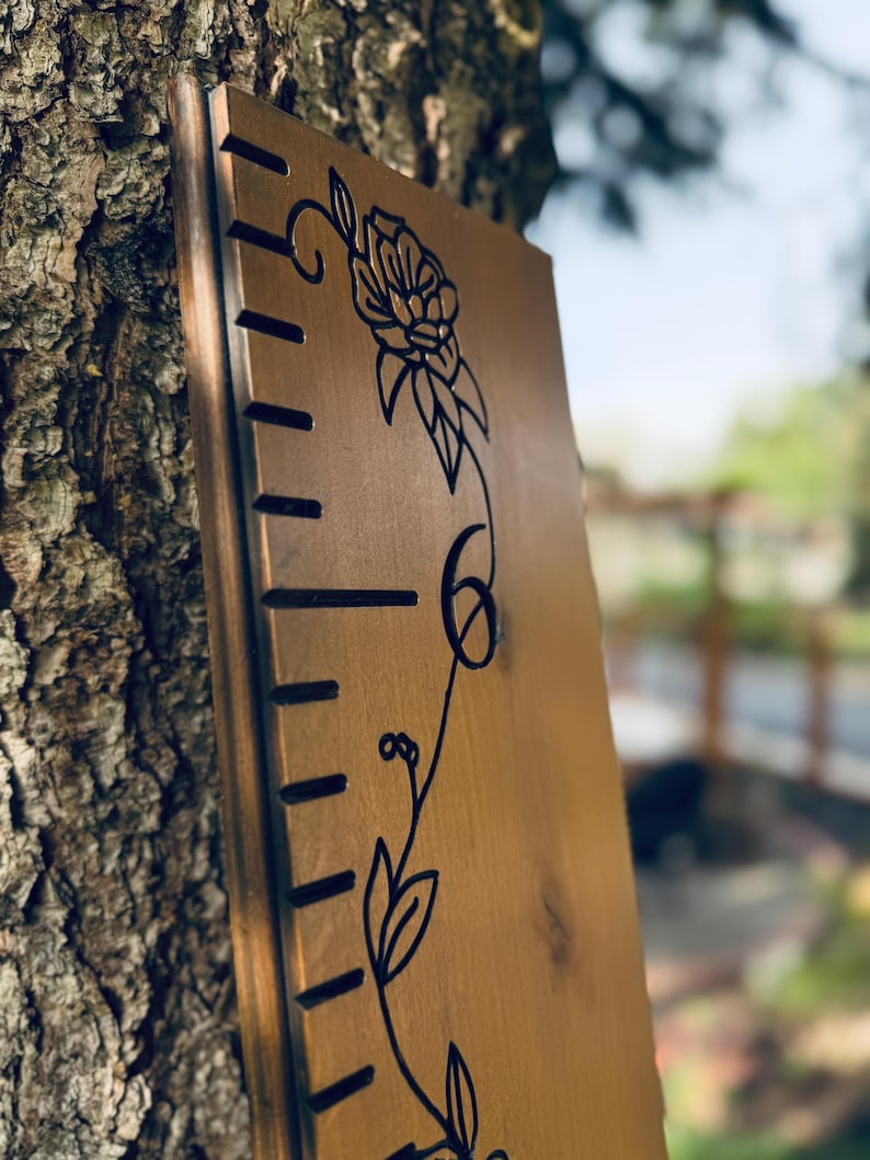 Op de afbeelding: Een verticaal houten groeimeterbord met een gladde, lichtbruine afwerking. Het bord is voorzien van zwarte, lasergegraveerde markeringen langs de linkerrand en een decoratieve zwarte lijntekening van een bloeiende bloemenrank in het midden.
