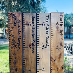 May include: A wooden growth chart with engraved names and height markings. The chart features the names "Charlie Morgan", "Jace Charles Wallace", "The Albee Family", and "The Gottlieb Family". The height markings are in feet and inches, up to 6 feet.