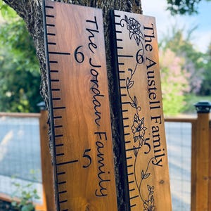 May include: Two vertical wooden growth charts with dark brown wood grain. The left board features the text The Jordan Family. The right board features the text The Austell Family with a floral vine illustration. Both have black measurement markings.