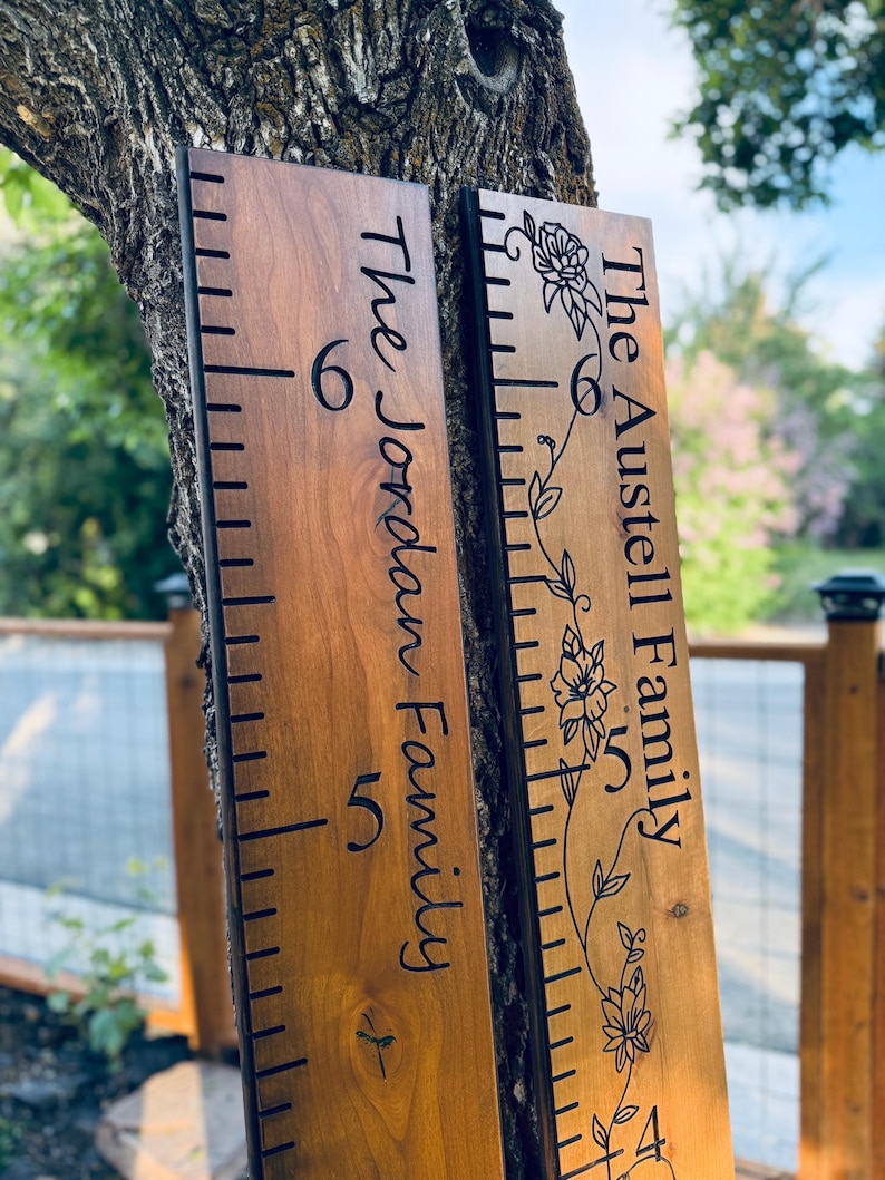May include: Two vertical wooden growth charts with dark brown wood grain. The left board features the text The Jordan Family. The right board features the text The Austell Family with a floral vine illustration. Both have black measurement markings.