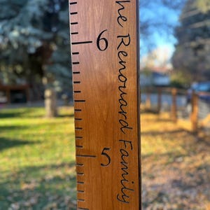 May include: A vertical wooden growth chart ruler with a medium brown finish. The board features engraved black measurement markings and the text The Renouard Family in a script font, with the numbers 5 and 6 clearly visible on the surface.