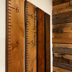 May include: Three vertical wooden growth charts featuring dark brown engraved measurement markings and numbers. Two boards display personalized text, specifically The Moodry Family and The Renouard Family, etched in a dark script font.
