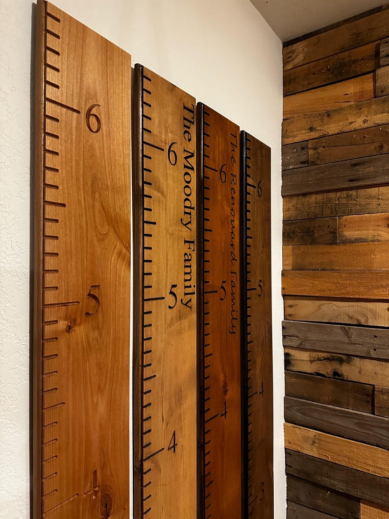 May include: Three vertical wooden growth charts featuring dark brown engraved measurement markings and numbers. Two boards display personalized text, specifically The Moodry Family and The Renouard Family, etched in a dark script font.