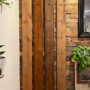 May include: Four vertical wooden growth charts with black measurement markings from 1 to 6 feet. Two boards feature custom text: The Renouard Family and The Moodry Family. The boards are finished in varying shades of light, medium, and dark brown wood.