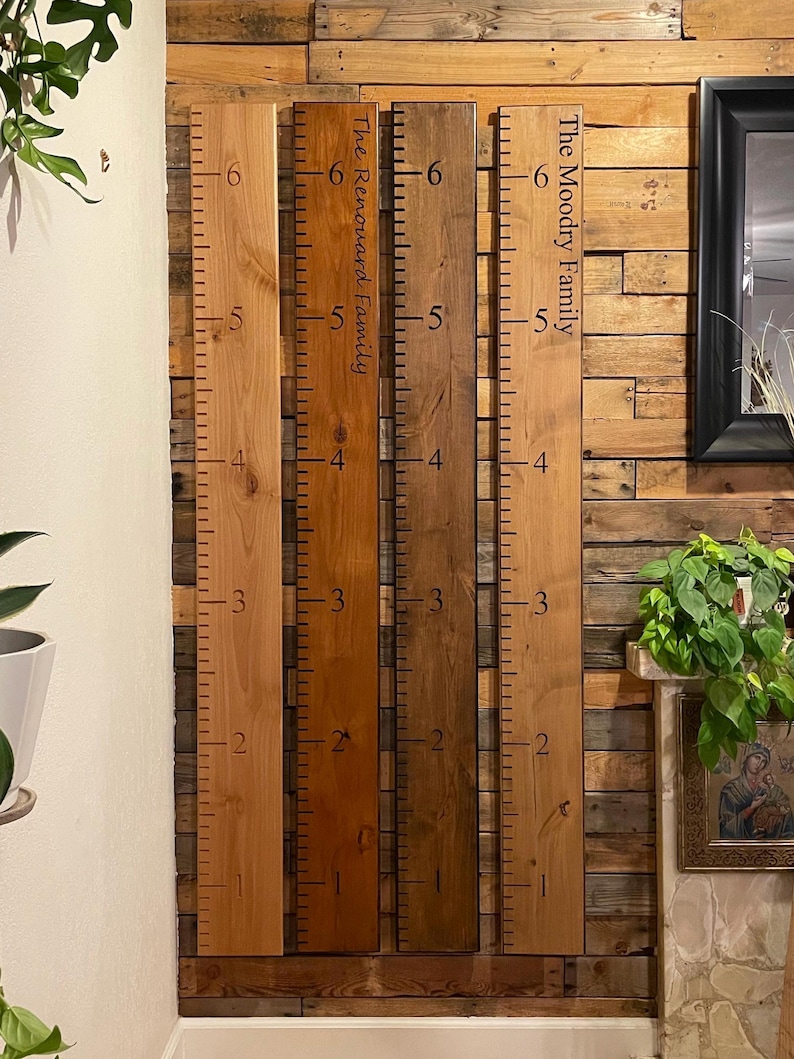 May include: Four vertical wooden growth charts with black measurement markings from 1 to 6 feet. Two boards feature custom text: The Renouard Family and The Moodry Family. The boards are finished in varying shades of light, medium, and dark brown wood.