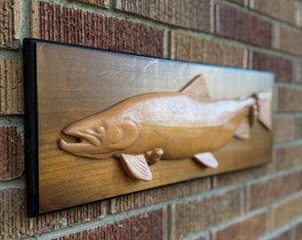May include: A wooden relief carving of a fish, mounted on a rectangular wooden plaque with a black border. The fish is a light brown color, with detailed fins and scales. The plaque is mounted on a brick wall.