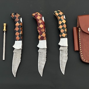 May include: Set of three folding knives with patterned wooden handles and serrated blades, alongside a sharpening rod and a brown leather sheath. The handles feature a mosaic design in shades of brown, red, and black.