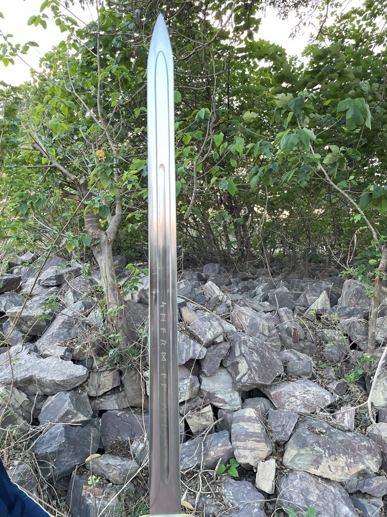 May include: A silver sword with a sharp point and a long, straight blade. The blade has markings on it that read "HFRM-F". The sword is standing upright in a pile of rocks.