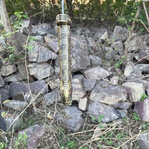 May include: A gold-colored sword hilt with intricate details and red gemstones. The hilt is hanging from a branch in a wooded area.