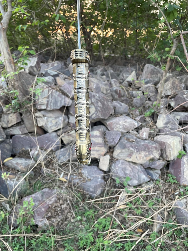 May include: A gold-colored sword hilt with intricate details and red gemstones. The hilt is hanging from a branch in a wooded area.